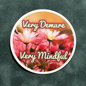 May include: A round sticker with a pink and white flower design. The sticker has the text "Very Demure" and "Very Mindful" in a yellow font.