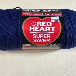May include: A skein of navy blue Red Heart Super Saver worsted medium weight yarn. The label features a red heart logo, the text 'Red Heart Super Saver', 'Worsted Medium 4', and 'Net Wt 7 oz / 198 g'. A small American flag icon is visible.
