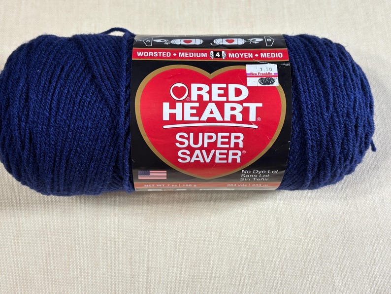 May include: A skein of navy blue Red Heart Super Saver worsted medium weight yarn. The label features a red heart logo, the text 'Red Heart Super Saver', 'Worsted Medium 4', and 'Net Wt 7 oz / 198 g'. A small American flag icon is visible.