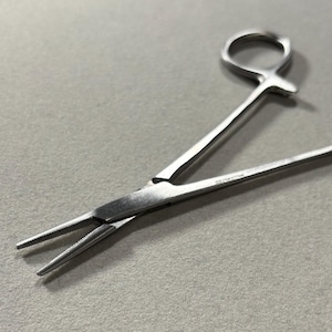 May include: A stainless steel hemostat with a locking mechanism, featuring two finger rings at one end and thin, serrated, pointed tips at the other. The metal instrument has a polished finish and a hinge that allows the jaws to open and close.