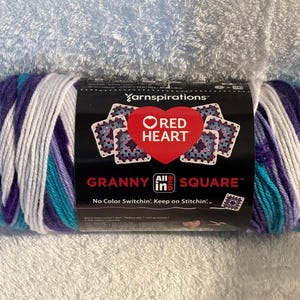 Pode incluir: Um novelo de fio Red Heart All in One Granny Square com uma paleta de cores variadas em branco, roxo claro, roxo escuro e azul-petr&oacute;leo. O r&oacute;tulo preto exibe o log&oacute;tipo da marca, um cora&ccedil;&atilde;o vermelho e o texto Granny Square.