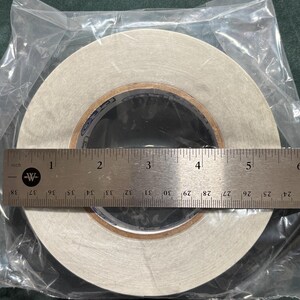 May include: A roll of white adhesive tape encased in a clear plastic bag, positioned behind a stainless steel ruler. The ruler shows the tape roll has a diameter of approximately 5.5 inches. The inner core of the tape roll features blue text.