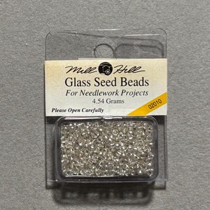 May include: A clear plastic package containing Mill Hill Glass Seed Beads for needlework projects. The label displays the text Mill Hill Glass Seed Beads For Needlework Projects 4.54 Grams Please Open Carefully and the number 02010.