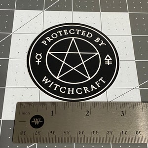 May include: A circular black sticker with a white border featuring a central pentagram, the text "PROTECTED BY WITCHCRAFT," and two alchemical symbols. A metal ruler placed below the sticker shows it has a diameter of approximately 3 inches.