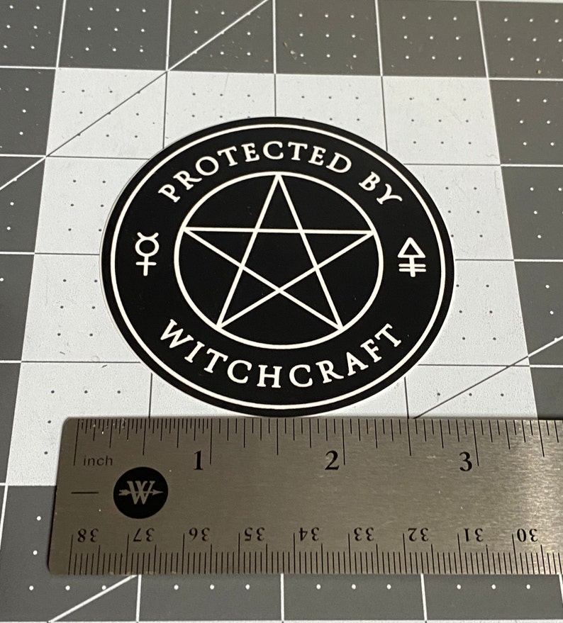 May include: A circular black sticker with a white border featuring a central pentagram, the text "PROTECTED BY WITCHCRAFT," and two alchemical symbols. A metal ruler placed below the sticker shows it has a diameter of approximately 3 inches.