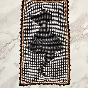 May include: A rectangular black crochet doily featuring a central silhouette of a ballerina in a tutu. The piece is constructed with a grid-like mesh pattern and is finished with a decorative black and orange scalloped border edge.