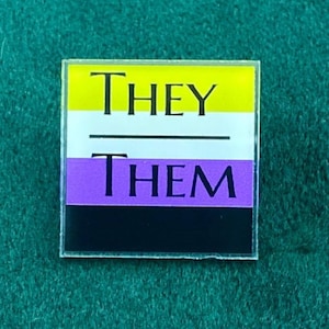 May include: A square pin with the words "THEY" and "THEM" in black, separated by a line. The pin features the non-binary pride flag colors: yellow, white, purple, and black.