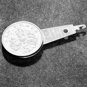 May include: A silver-colored metal tool featuring a circular end resembling a 5 New Pence coin with a crown and thistle design, attached to a flat, tapered arm with a small circular hole and a hook-shaped tip at the far end of the object.