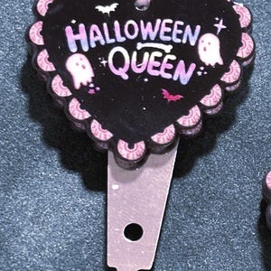 May include: A heart-shaped Halloween-themed hair clip. The black clip features the words "HALLOWEEN QUEEN" with ghost and bat graphics. The edges are scalloped with pink and white accents, and the clip is silver.