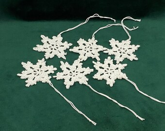 Op de afbeelding: Een set van vijf witte gehaakte sneeuwvlokornamenten, elk met een zespuntig sterontwerp met ingewikkelde kantachtige patronen en een dun wit touwtje aan de bovenkant om ze op te hangen, gerangschikt in een groep op een donkere ondergrond.