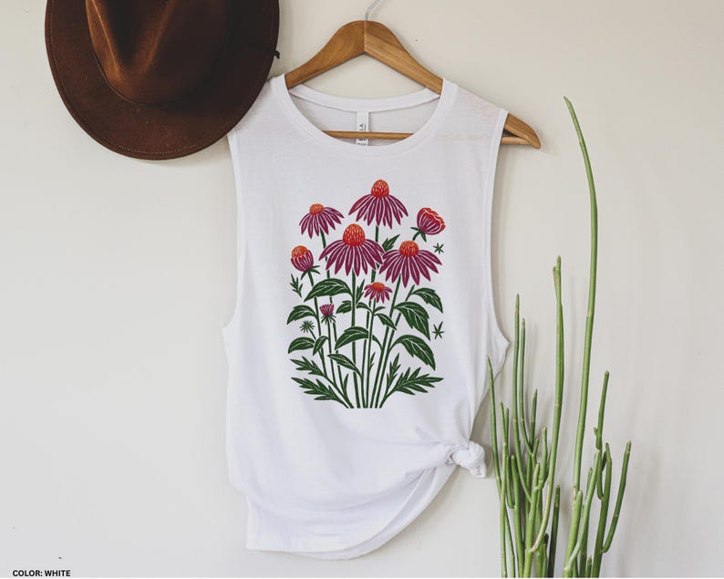 May include: White sleeveless shirt with a floral design. The graphic features purple coneflowers with orange centers and green leaves. The shirt is hanging on a wooden hanger against a white wall.