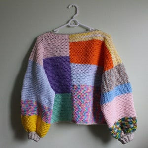 May include: A colourful crocheted jumper with a patchwork design. The jumper has a light blue, pink, purple, green, orange, and yellow colour scheme. The jumper has long sleeves and a relaxed fit.