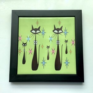 May include: Framed green tile with a retro design featuring five black cats with white eyes and pink and blue accents. The cats are surrounded by stylized stars.