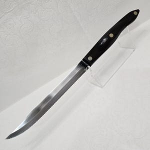 Op de afbeelding: Een gekarteld steakmes met een zilverkleurig lemmet en een zwarte handgreep. De handgreep is bevestigd met drie zilveren klinknagels. Het mes wordt gepresenteerd op een transparante acrylstandaard. Het lemmet is ongeveer 20 cm lang.