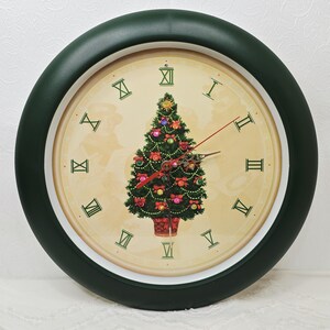 May include: A round wall clock with a dark green frame and a cream-colored face. The dial features green Roman numerals and a central illustration of a decorated Christmas tree with red bows and colorful ornaments, plus three thin clock hands.