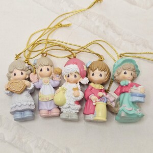 May include: A set of five porcelain-style figurines designed as hanging ornaments, each featuring a child with teardrop-shaped eyes. The figures wear pastel-coloured clothing and hold various items including a heart-shaped cookie, a bag, and a basket.