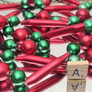 May include: A festive garland composed of alternating red and green glass beads and elongated cylindrical ornaments. The beads and cylinders feature a smooth, reflective finish. A small wooden square tile with the letter A and number 1 is visible.