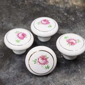 May include: Four white ceramic cabinet knobs with a floral rose design. Each knob features a pink rose with green leaves, encircled by a gold or silver metallic trim. The knobs are set against a dark, textured surface.