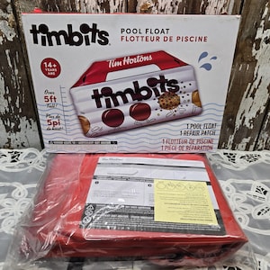 Pode incluir: Um flutuador de piscina Tim Hortons numa caixa, com o texto "Timbits" e "Flutuador de piscina". A caixa também inclui o texto "Mais de 5 pés de altura!" e "14+ anos". Um flutuador de piscina vermelho está num saco plástico.