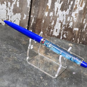 Könnte beinhalten: Blauer Stift mit silbernem Clip und dekorativem Schaft mit Landschaftsmotiv. Der Stift wird auf einem transparenten Acrylständer präsentiert, der seine leuchtende Farbe und seine komplizierten Details hervorhebt. Der Stift ist etwa 14 cm lang.
