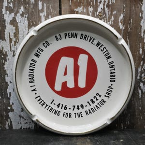 Op de afbeelding: Witte asbak met een rode cirkel in het midden met de witte tekst "A1". De asbak heeft een gouden rand en de tekst "A1 RADIATOR MFG CO. EVERYTHING FOR THE RADIATOR SHOP" rondom.