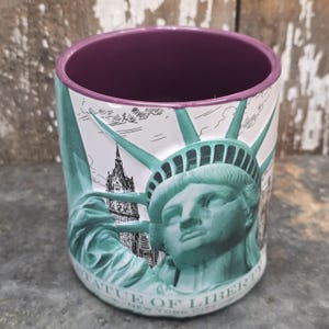 Könnte beinhalten: Eine Keramik-Tasse mit einem Design der Freiheitsstatue in Türkis und Weiß, mit einem lila Innenraum. Auf der Tasse steht der Schriftzug "Statue of Liberty New York City". Die Tasse ist zylindrisch und hat eine detaillierte Illustration der Statue.