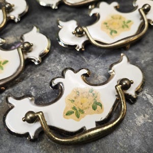 Op de afbeelding: Verschillende witte en gouden ladehandgrepen met een bloemenontwerp. Elke handgreep heeft een witte emaille basis met een geel en groen bloemenmotief. De handgrepen hebben een goudkleurige handgreep en afwerking, en een kleine, ronde knop.