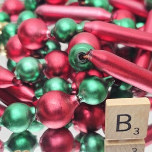 May include: A garland of red and green glass beads featuring alternating spherical and elongated cylindrical shapes with a matte finish. A small wooden square tile with the black letter B and the number 3 is positioned in the lower right corner.