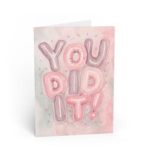 May include: A greeting card featuring the text YOU DID IT! rendered in balloon-style lettering. The balloons are coloured in shades of soft pink and muted mauve against a watercolour background of blended pink and grey tones with small star accents.