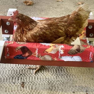May include: A red wooden frame with white wheels and a red fabric cushion with a chicken print. A brown chicken is sitting on the cushion.