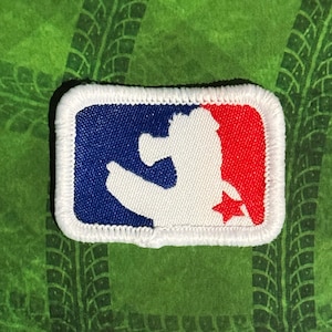 May include: Embroidered patch with a white border. The patch features a silhouette of a person in a baseball stance, with a blue background on the left and a red background on the right. A white star is visible on the red side.