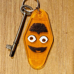 May include: A vintage-style key and a yellow motel keychain with a smiling face design. The keychain has a black mustache, eyebrows, and white eyes. The key is made of metal.