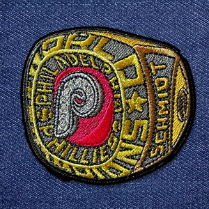 May include: Embroidered patch featuring the Philadelphia Phillies logo in red, white, and gray, with gold lettering and accents. The patch includes the name "SCHMIDT" and a star. The patch is on a blue denim background.