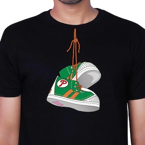 Könnte beinhalten: Ein schwarzes T-Shirt mit kurzen Ärmeln und einer Grafik von zwei grünen Turnschuhen mit orangefarbenen Streifen und einem roten Buchstaben P an der Seite. Die Turnschuhe sind mit ihren orangefarbenen Schnürsenkeln verbunden und hängen vertikal.