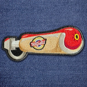 May include: Embroidered patch featuring a stylized hot dog with a red bun, yellow mustard, and the "Hatfield" logo. The patch has a white, black, and gold border and is set against a blue denim background.