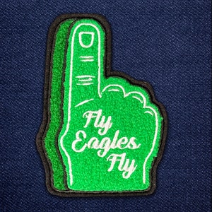 May include: A green foam finger patch with the words "Fly Eagles Fly" in white script. The patch has a black border and is set against a blue denim background. The foam finger is a classic sports fan accessory.