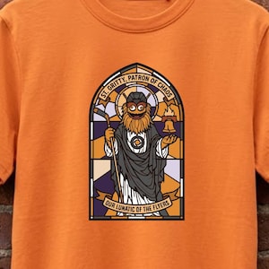 Könnte beinhalten: Ein orangefarbenes T-Shirt mit kurzen Ärmeln und einem Grafikdruck eines bärtigen Charakters mit Eishockeyschläger in einem Buntglasfenster-Rahmen. Der Text lautet ST. GRITTY, PATRON OF CHAOS und OUR LUNATIC OF THE FLYERS.