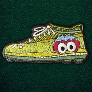 May include: Embroidered patch in the shape of a running shoe. The shoe is primarily yellow-green with white laces and a cartoonish face with large eyes. The patch has a black border and is set against a dark green background.