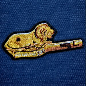 May include: Embroidered patch featuring a golden lion with a brown mane and the text "PHILADELPHIA ZOO" in gold. The patch is outlined in black and has a brown and gold handle-like shape.