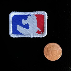 May include: A rectangular patch featuring the MLB logo in red, white, and blue. The patch is bordered with white stitching. A US one-cent coin is next to the patch for size comparison.