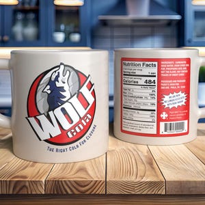 Puede incluir: Dos tazas de cerámica blanca. Una muestra el logo "WOLF COLA", un lobo aullando en un círculo rojo y blanco. La otra muestra información nutricional. Sobre una superficie de madera.