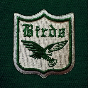 May include: Embroidered patch featuring a shield design with the word "Birds" in green gothic lettering. A green eagle graphic is centered below the text. The patch has a white and green border, set against a dark green fabric background.