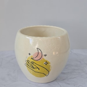 May include: Cream-colored ceramic pot with a hand-drawn design. The design features a hand, crescent moon, and stars over a yellow circle. The pot has a smooth, glossy finish and a wide opening. Ideal for holding plants or as a decorative item.