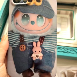 May include: A phone case featuring a cartoon character with a blue denim hat and overalls. The character has a large, smiling face with light blue eyes. A small pink bunny with a heart is attached to the overalls.