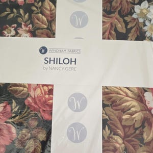 Op de afbeelding: Een stoffenbundel uit de Shiloh-collectie van Nancy Gere voor Windham Fabrics. De stof heeft een donkere achtergrond met een bloemenpatroon in roze, crème en bruine tinten. Een witte papieren band met het merklogo en tekst ligt over de stof heen.
