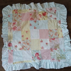 May include: A light blue baby quilt with a patchwork design featuring floral patterns in shades of pink, yellow, and green. The quilt has a ruffled edge.