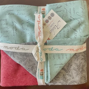 May include: A bundle of fabric squares in shades of teal, gray, and red, tied with a cream ribbon printed with "moda". The fabric features script and floral patterns. A tag reads "Atelier".