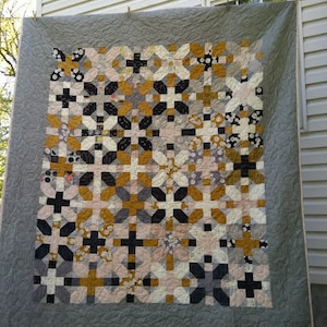 Könnte beinhalten: Eine handgefertigte Steppdecke mit einem geometrischen Muster aus Kreuzen und floralen Formen in Schwarz-, Gold-, Grau- und Cremetönen. Die Steppdecke hat einen grauen Rand und hängt draußen.