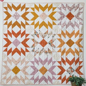 Könnte beinhalten: Eine große, gesteppte Decke mit einem Sternmuster. Der Quilt hat ein Patchwork-Design mit verschiedenen Farben, darunter Weiß, Orange, Gelb und Rosa. Die Sternformen sind in einem Rastermuster angeordnet und schaffen ein optisch ansprechendes und gemütliches Textil.