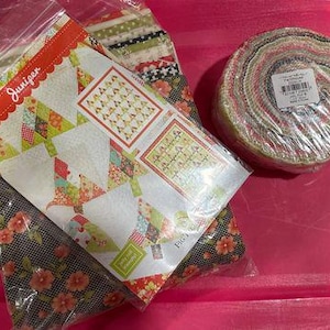 Puede incluir: Un paquete de tela con un patrón de colcha con un diseño verde, amarillo y rosa. El paquete incluye un panel de tela con el texto "Juniper" y un rollo de tela con un patrón multicolor.